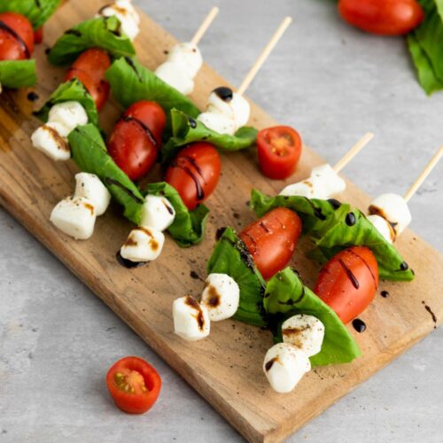 Caprese appetizers on a wooden board, with cherry tomatoes, mozzarella balls, and basil leaves, drizzled with balsamic glaze.