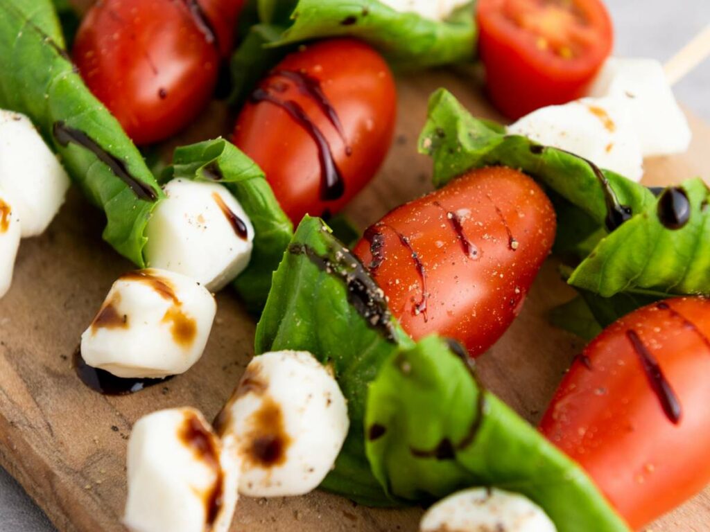 Caprese appetizer with cherry tomatoes, mozzarella, basil leaves, and balsamic glaze on a wooden board.