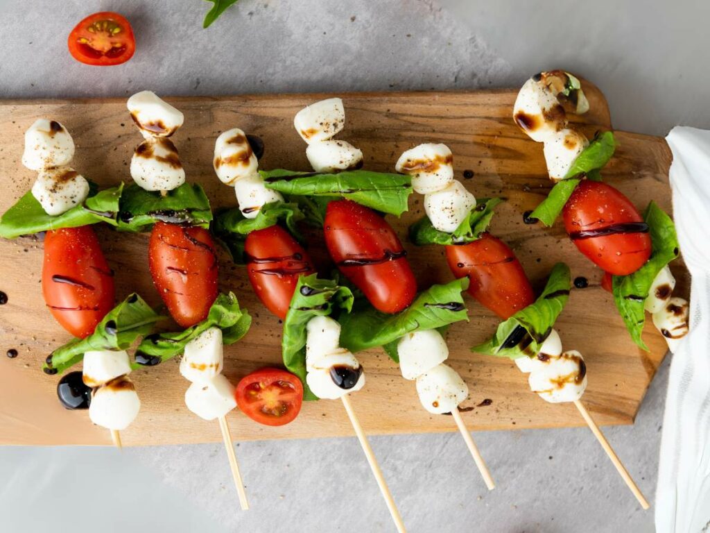 Four caprese appetizers with tomatoes, mozzarella, and basil on a wooden board, drizzled with balsamic glaze.