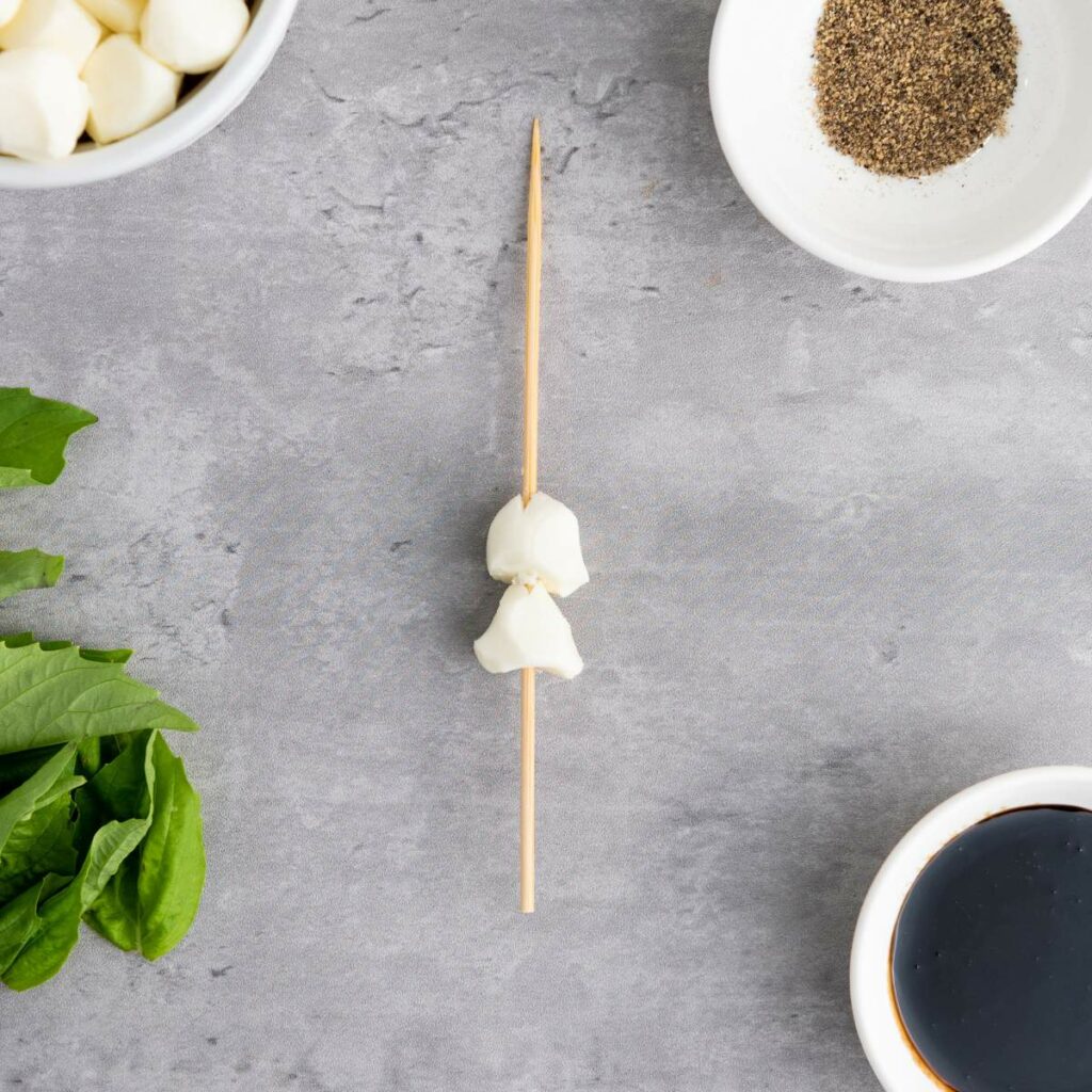 A skewer with two mozzarella balls on a gray surface, surrounded by bowls of black pepper, balsamic vinegar, and leafy greens.
