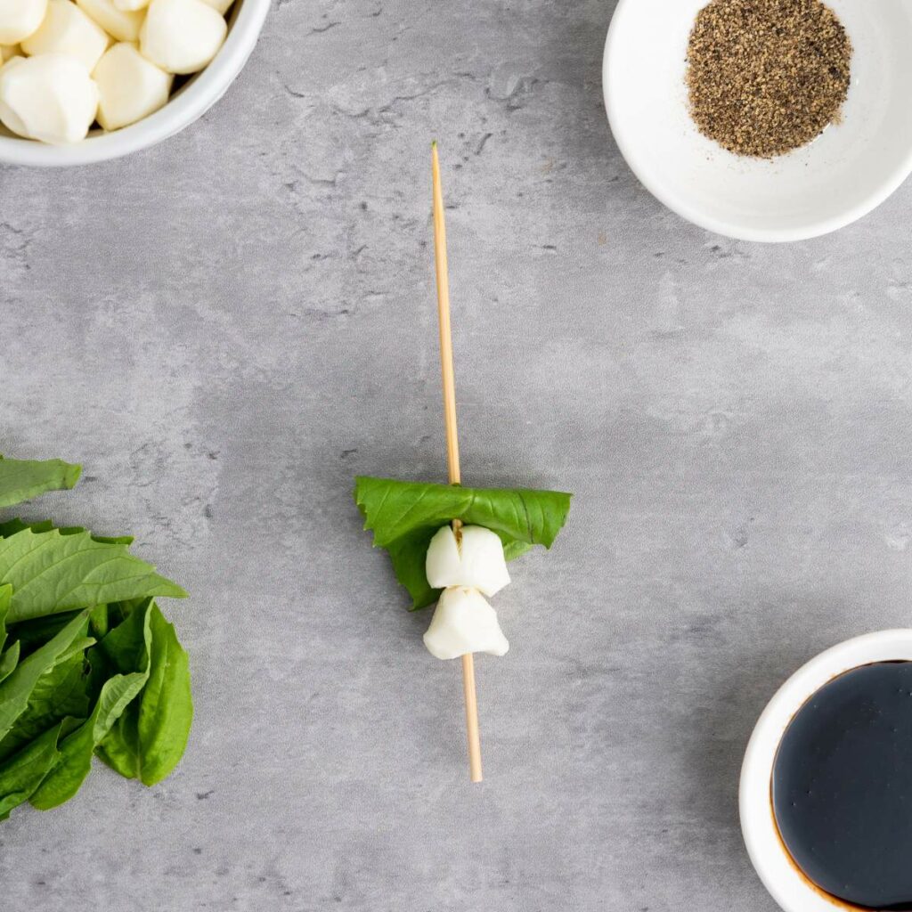A skewer with basil leaves and mozzarella is on a gray surface.
