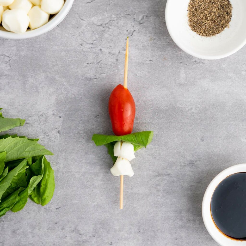 Wooden skewer with mozzarella, basil, and cherry tomato on a gray surface.