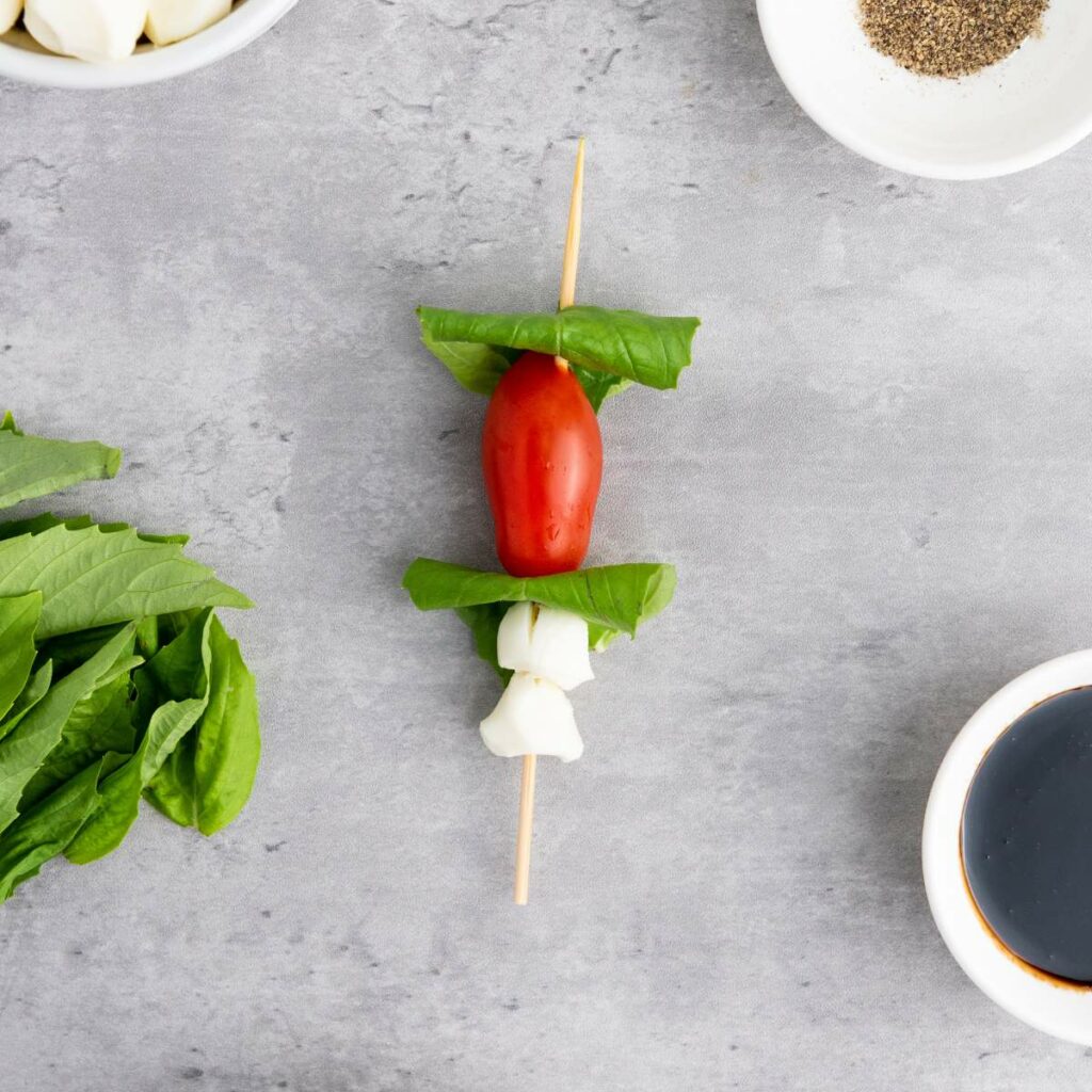 Skewer with a cherry tomato, basil leaves, and mozzarella, on a gray surface.
