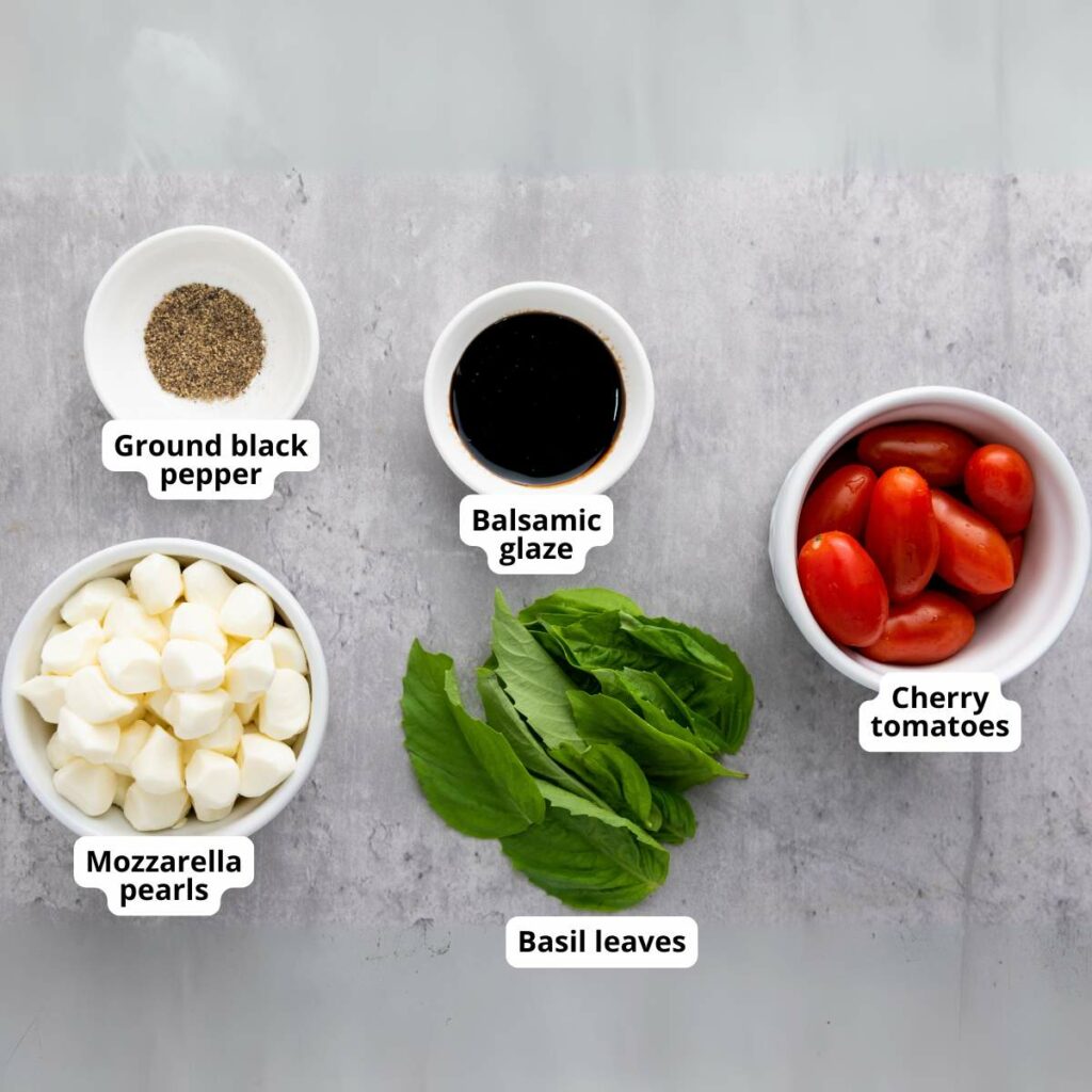 Five bowls with ingredients on a gray surface: ground black pepper, balsamic glaze, cherry tomatoes, mozzarella pearls, and basil leaves.