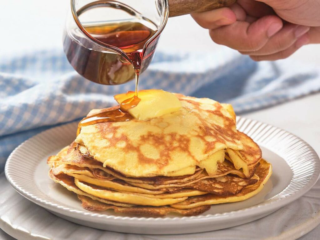 A stack of pancakes on a white plate with a pat of butter. Syrup is being poured over the pancakes from a small glass jug. A blue and white cloth is in the background.