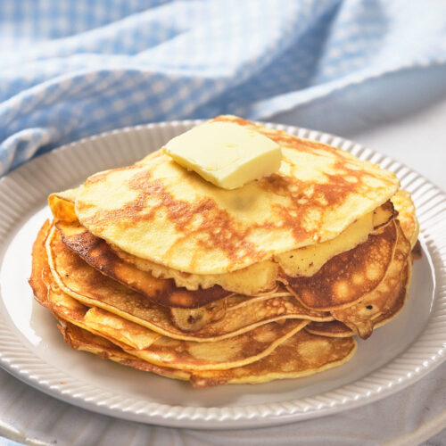 A stack of pancakes topped with a pat of butter on a white plate, placed on a light-colored tablecloth.