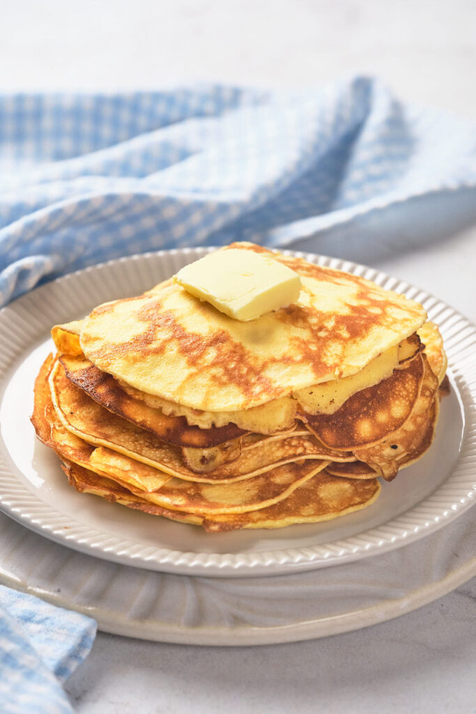 A stack of pancakes topped with a pat of butter on a white plate, placed on a light-colored tablecloth.