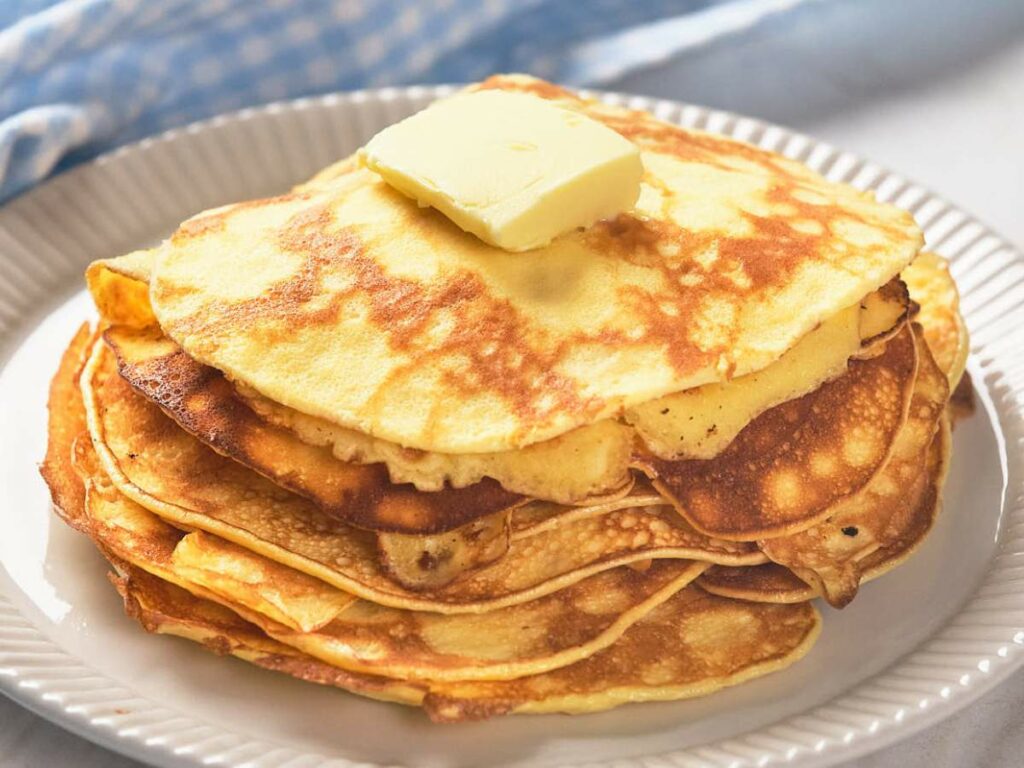 A stack of golden pancakes topped with a pat of butter on a white plate, placed on a light blue and white checkered cloth.