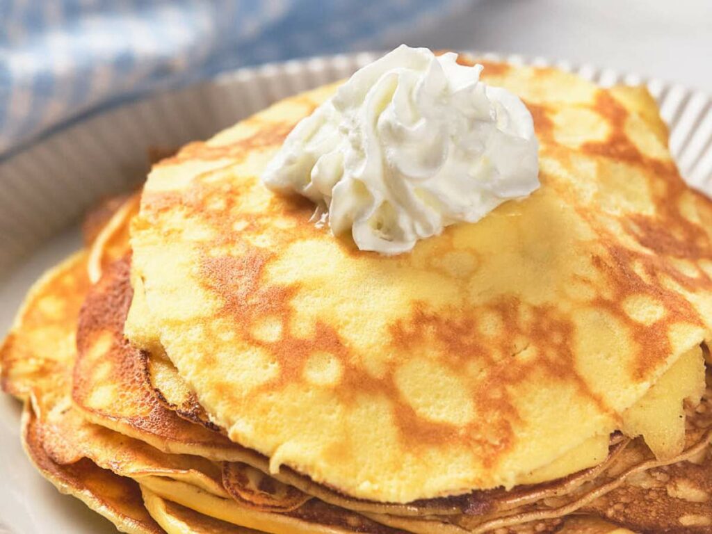 A stack of pancakes topped with whipped cream on a plate, with a blue checkered cloth in the background.