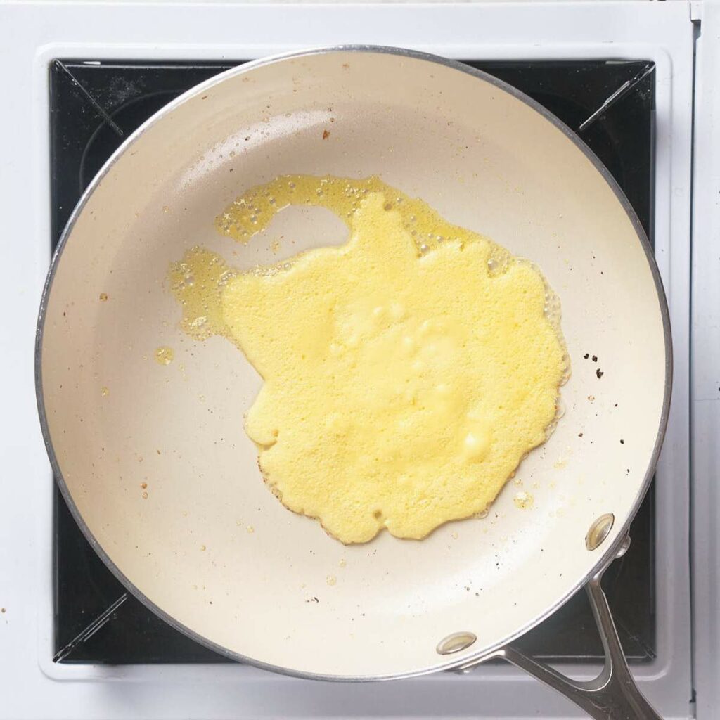 A partially cooked omelet spreads unevenly in a white frying pan on a stovetop.
