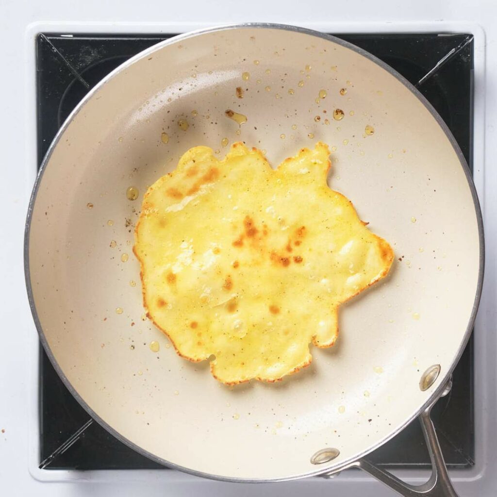 A pancake cooking in a light-colored frying pan on a stovetop.