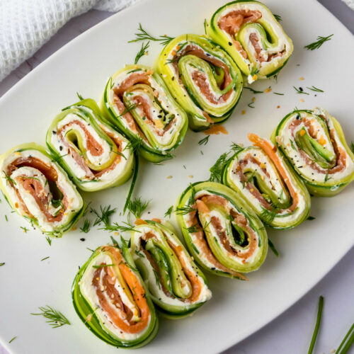 Sliced cucumber roll ups filled with smoked salmon, cream cheese, and herbs, arranged neatly on a white plate with scattered dill garnish.