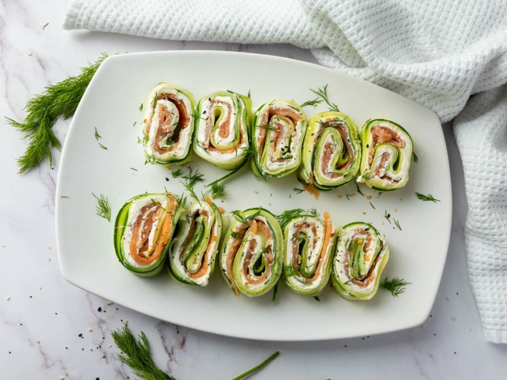 Sliced cucumber roll ups filled with cream cheese, smoked salmon, and herbs, arranged neatly on a white rectangular plate.