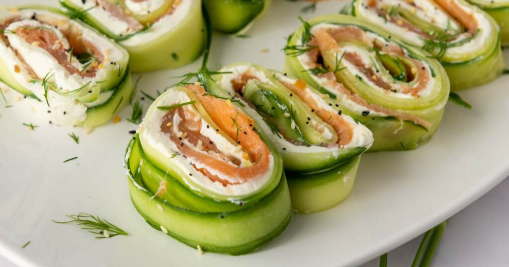 Cucumber roll ups filled with cream cheese and smoked salmon, garnished with dill and black pepper, on a white plate.
