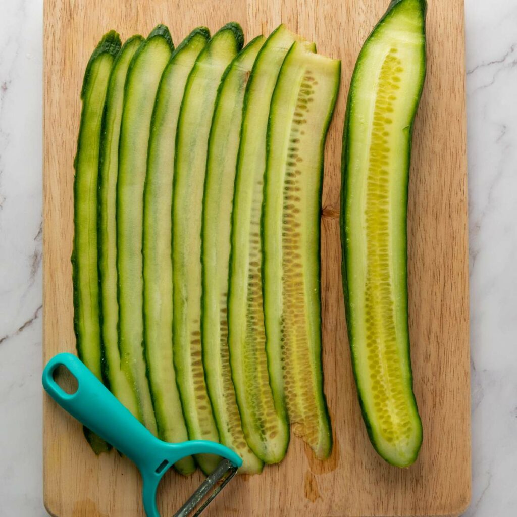 Thinly sliced cucumber on a wooden cutting board next to a teal vegetable peeler.