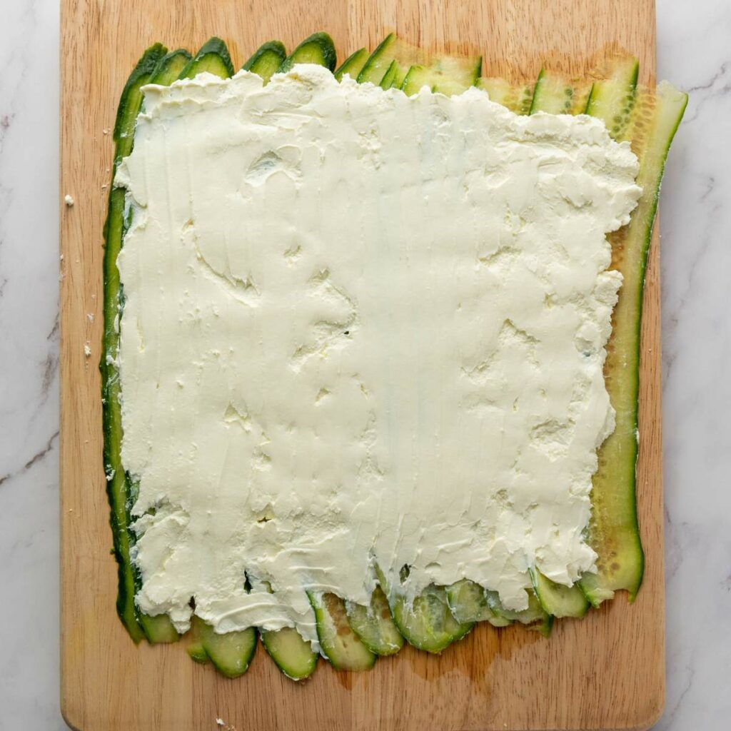 Sliced zucchini layered on a cutting board, topped with a spread of cream cheese.