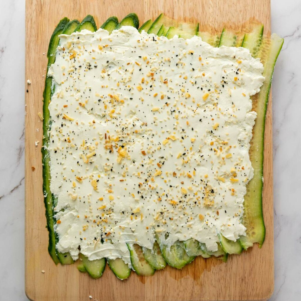 Cucumber slices on a wooden board topped with a layer of cream cheese and sprinkled with seasoning.