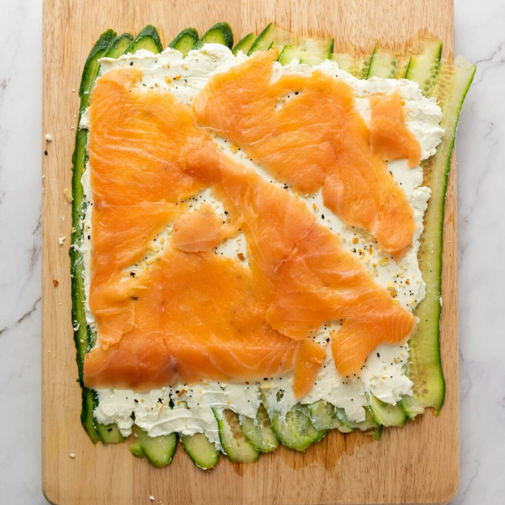 A wooden board with layered sliced cucumbers, cream cheese, and smoked salmon on top.