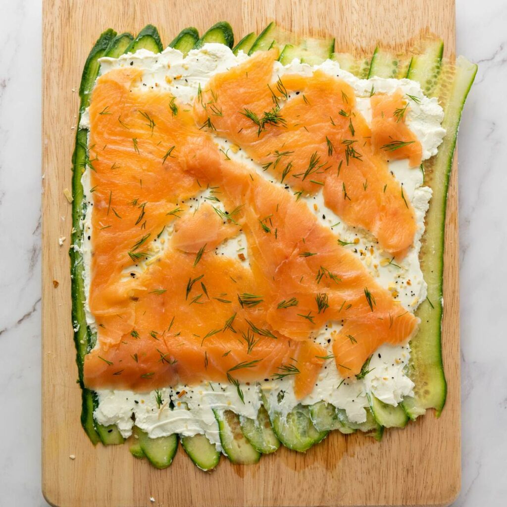 Cucumber slices, cream cheese, and smoked salmon are layered on a wooden board.