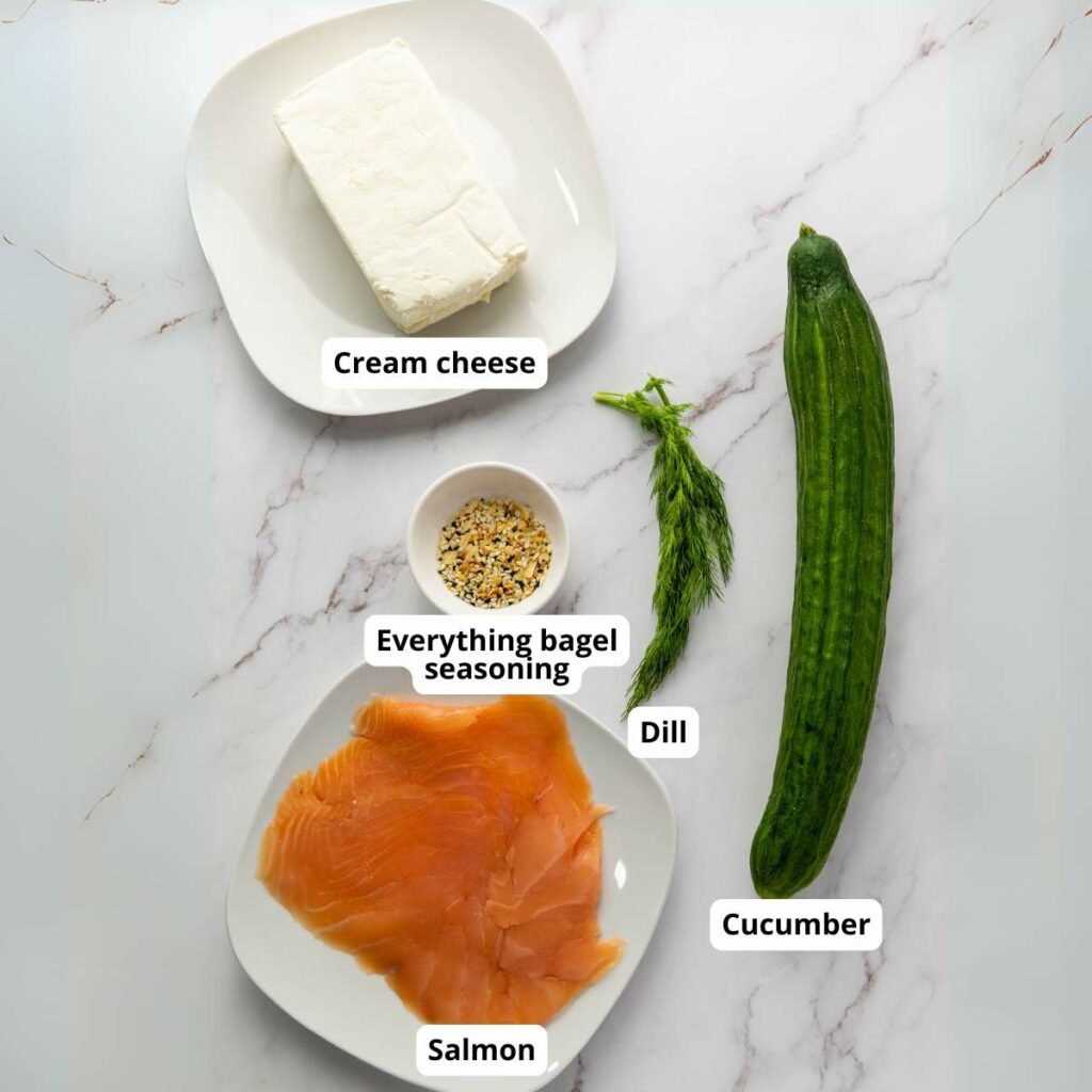 A marble surface with cream cheese, a cucumber, a sprig of dill, a plate of salmon, and a small bowl of everything bagel seasoning.