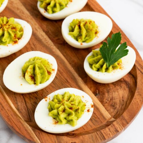 Deviled eggs with green filling on a wooden tray, garnished with paprika and parsley, placed on a marble surface.