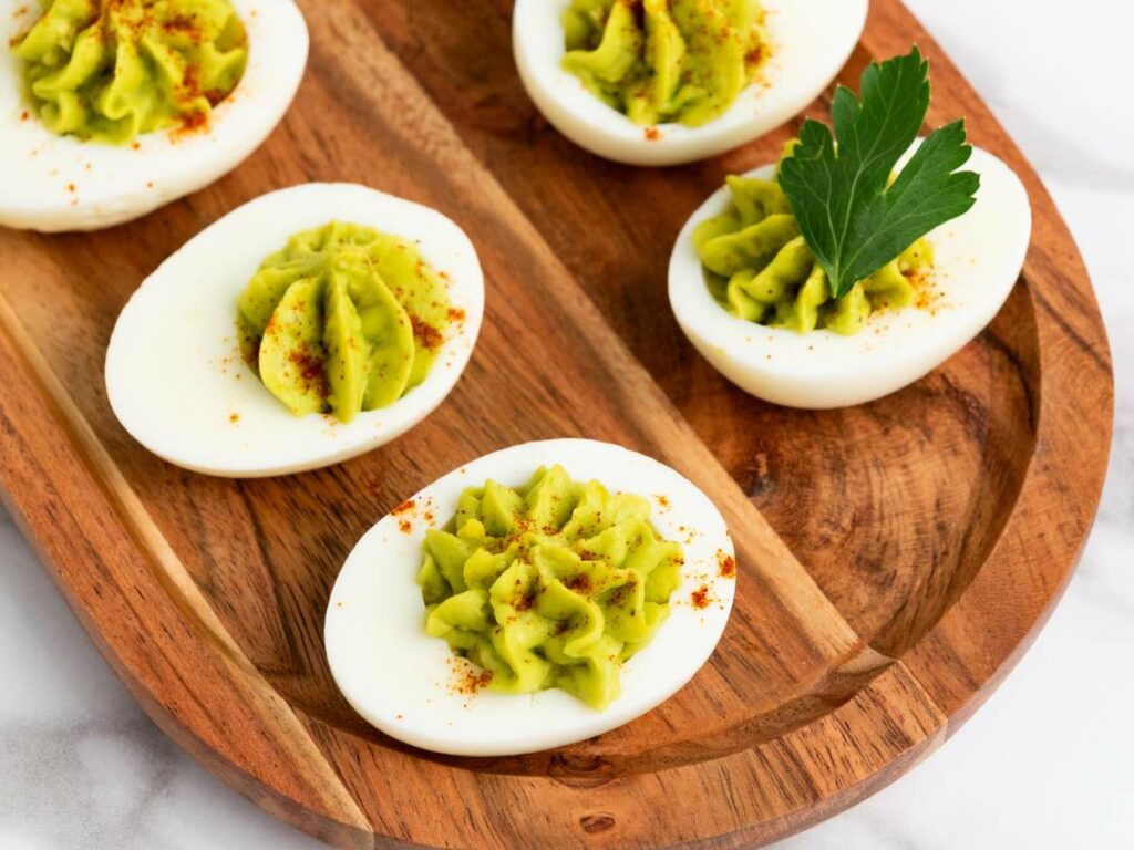 Deviled eggs with a green filling, sprinkled with paprika on a wooden tray. One egg is garnished with a parsley leaf.