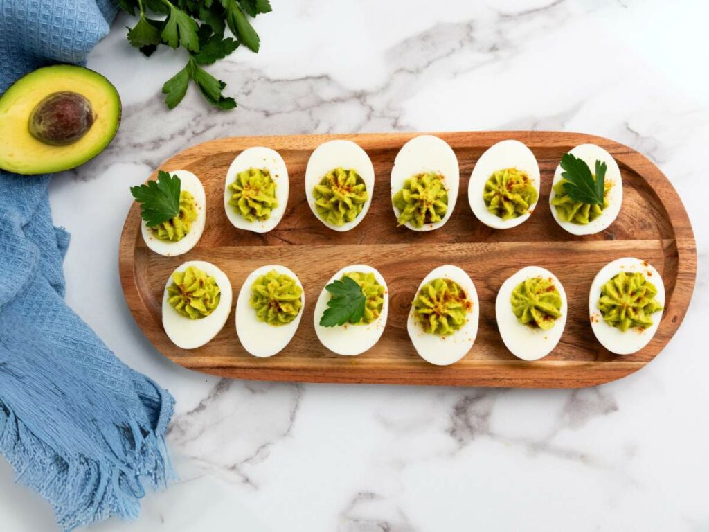 Deviled eggs topped with avocado filling and parsley on a wooden platter, placed on a marble surface with an avocado half and blue fabric nearby.