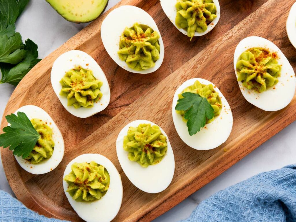 Deviled eggs garnished with green filling, a sprinkle of paprika, and parsley leaves are arranged on a wooden tray next to an avocado and blue cloth.