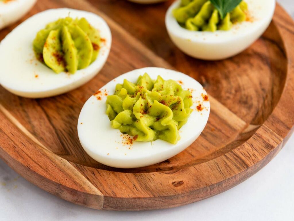 Halved deviled eggs with green filling garnished with paprika on a wooden platter.