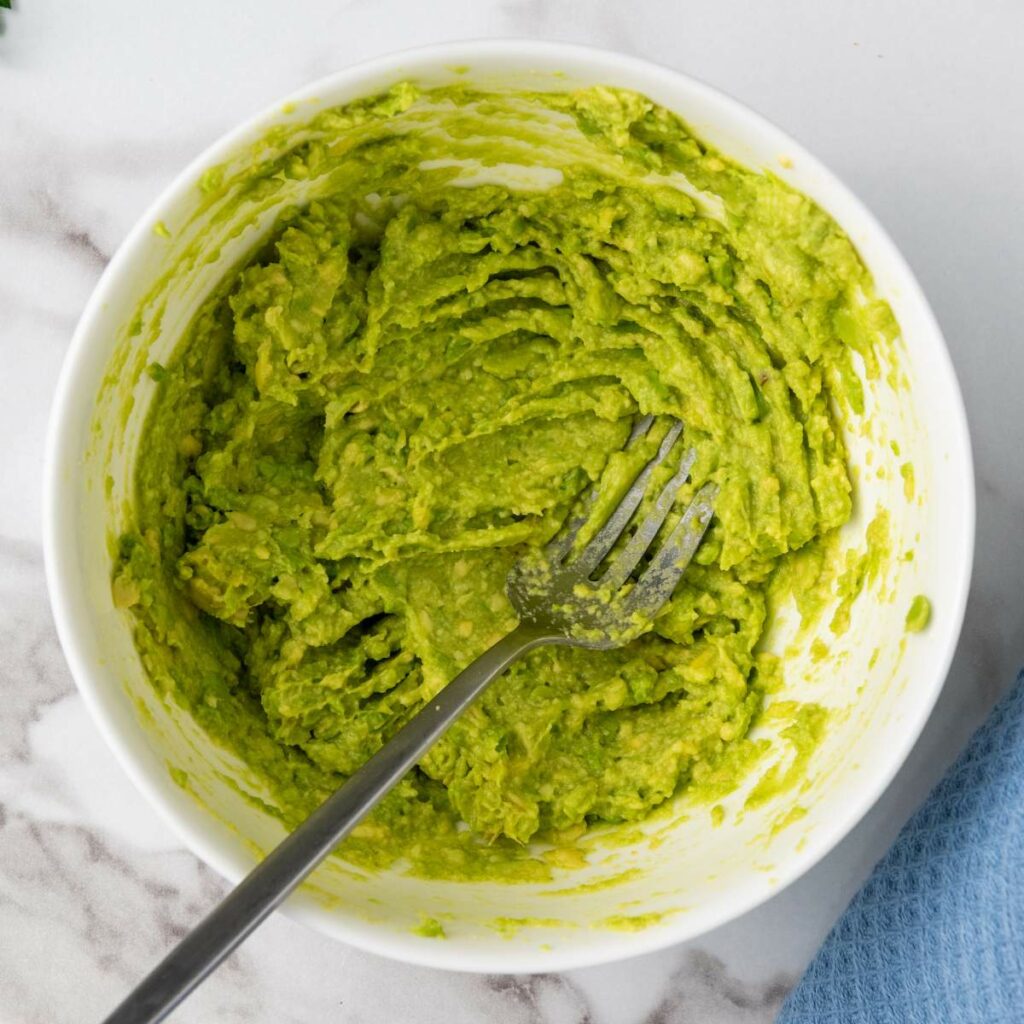A bowl of mashed avocado with a fork inside, placed on a marble surface next to a blue cloth.