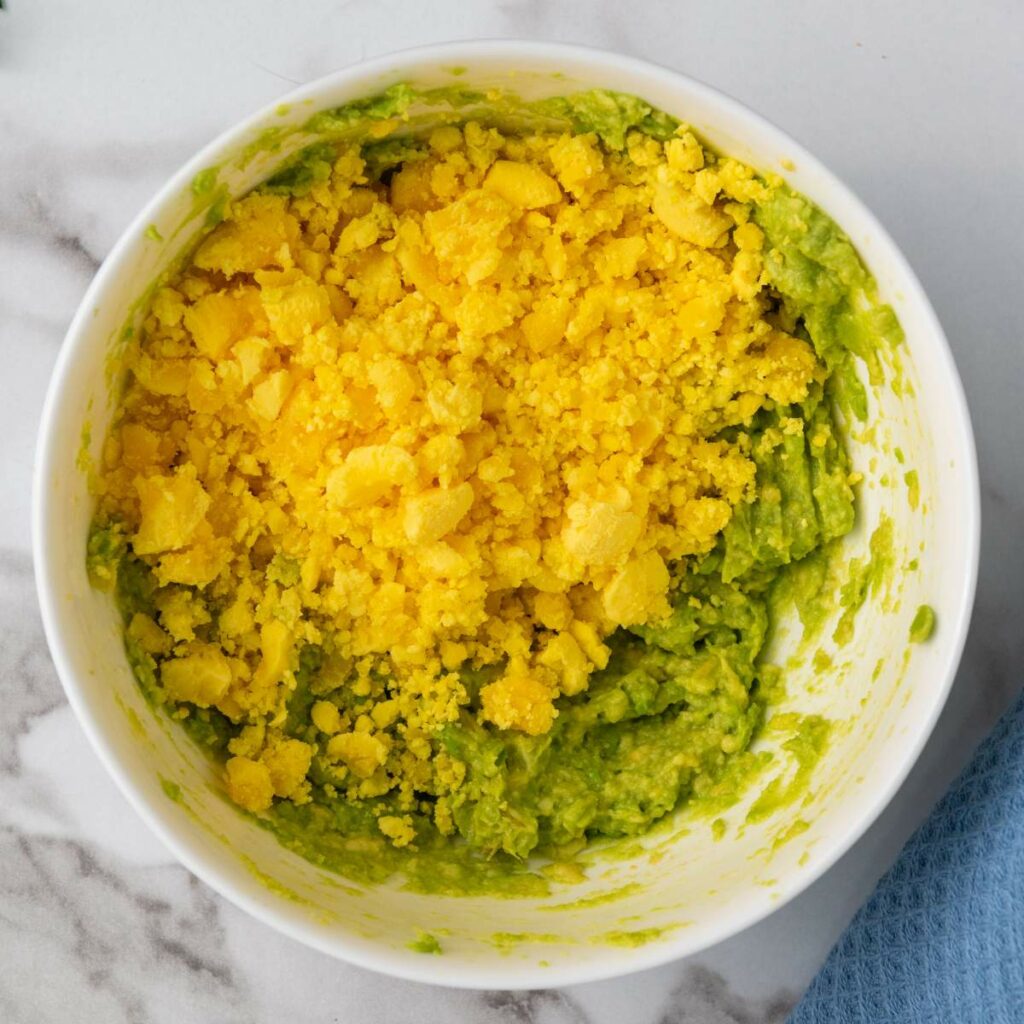 A bowl containing mashed avocado topped with crumbled yellow egg yolk on a marble surface.