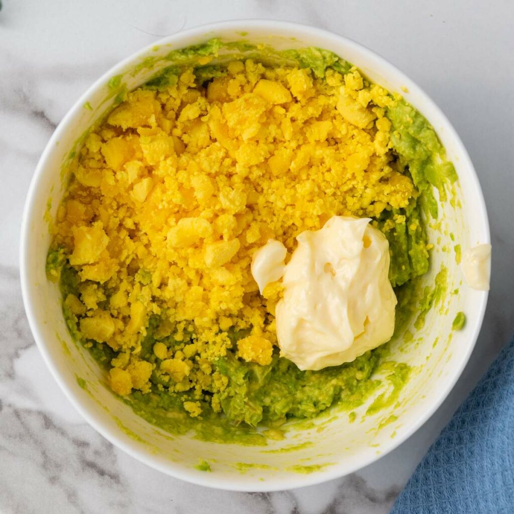 A bowl with mashed avocado, crumbled hard-boiled egg yolk, and a dollop of mayonnaise on a marble surface.