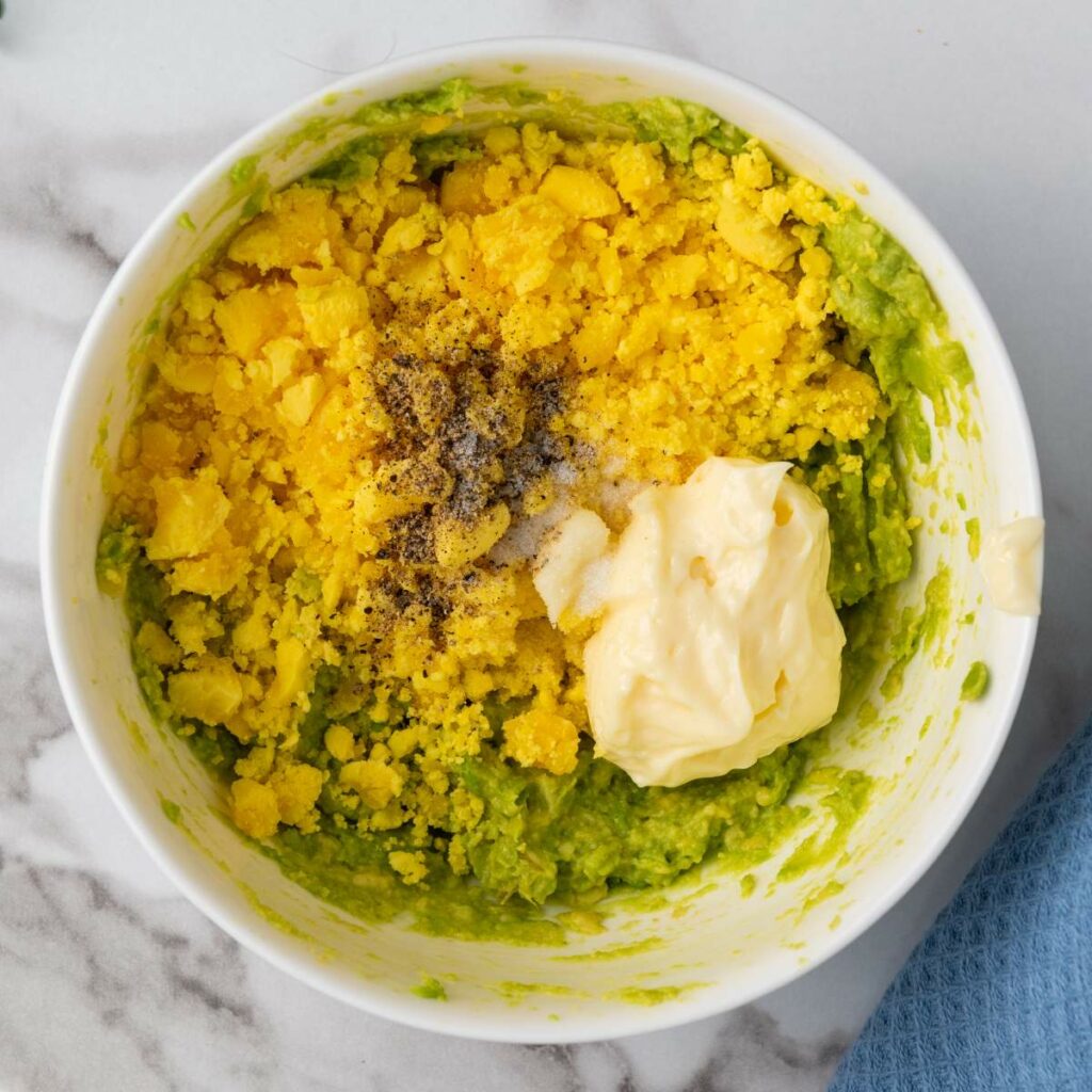 A bowl containing mashed avocado, crumbled egg yolks, a dollop of mayonnaise, salt, and pepper on a marble surface.