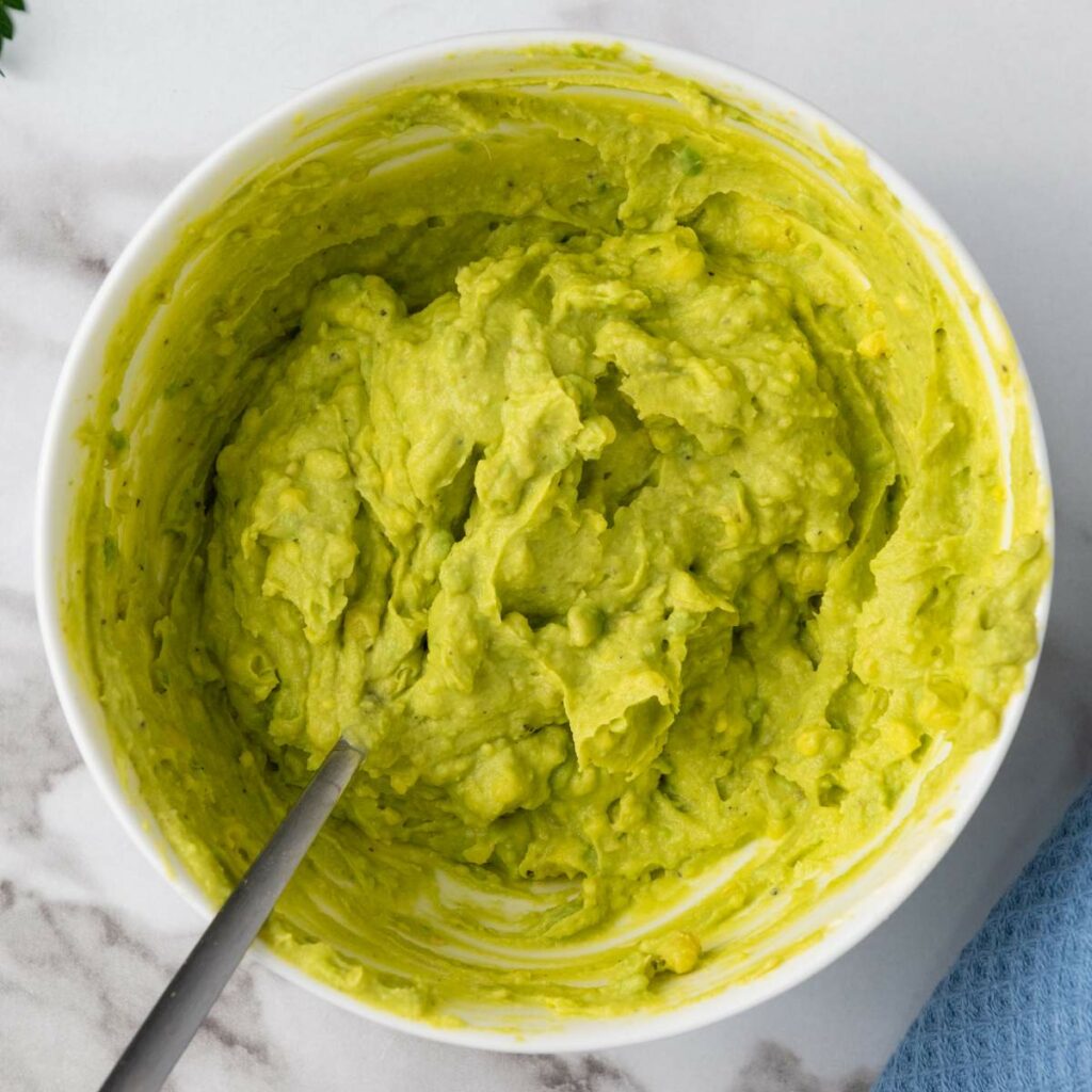 A bowl of mashed avocado with a fork, placed on a marble surface.