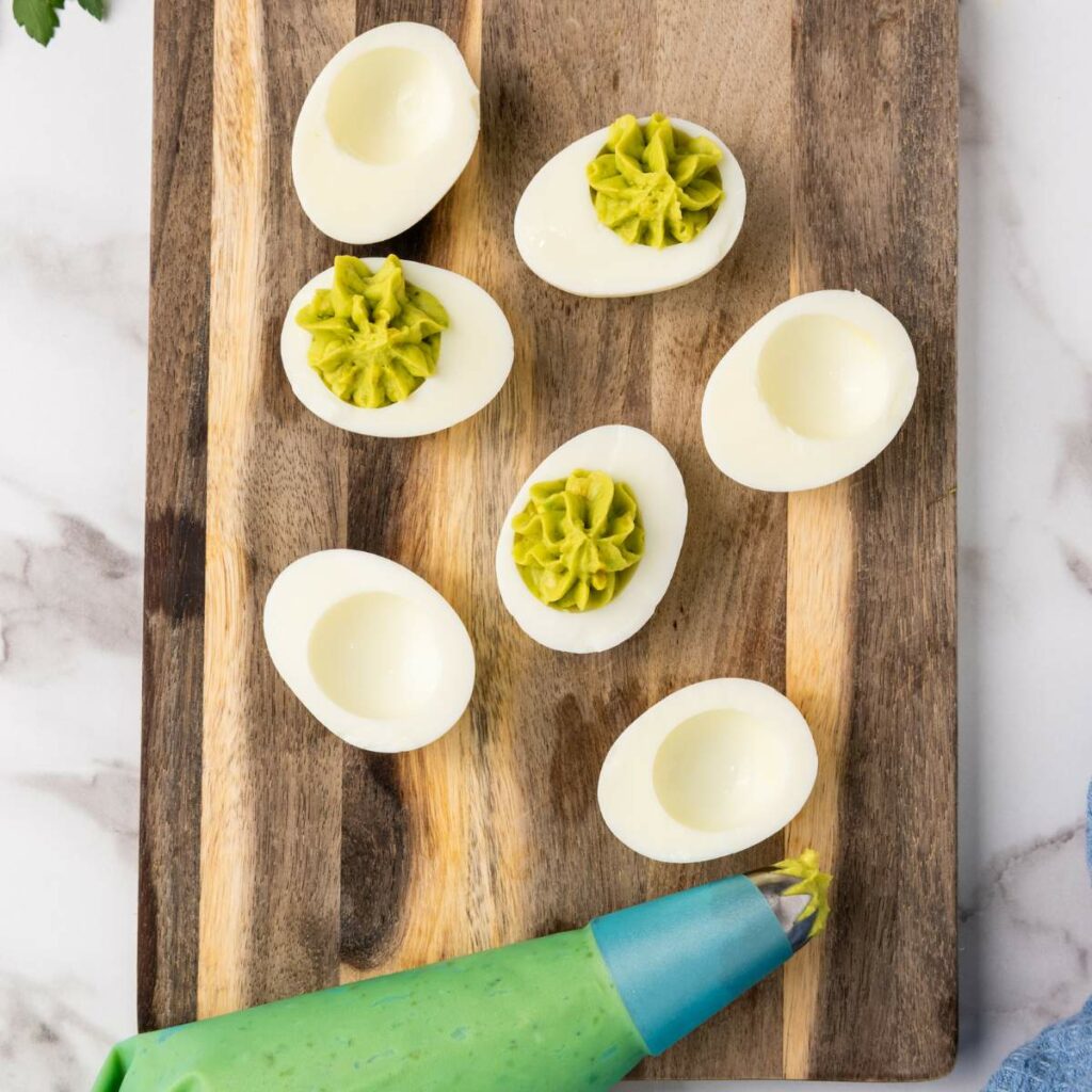 Six deviled eggs on a wooden board, with three filled with green avocado mixture.