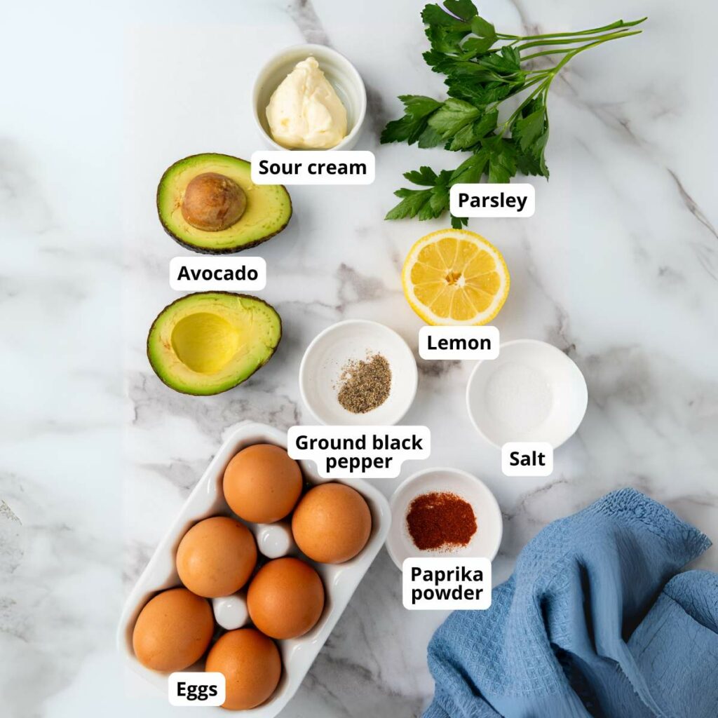 Ingredients on a marble surface: eggs, avocado, sour cream, parsley, lemon, ground black pepper, salt, paprika powder.