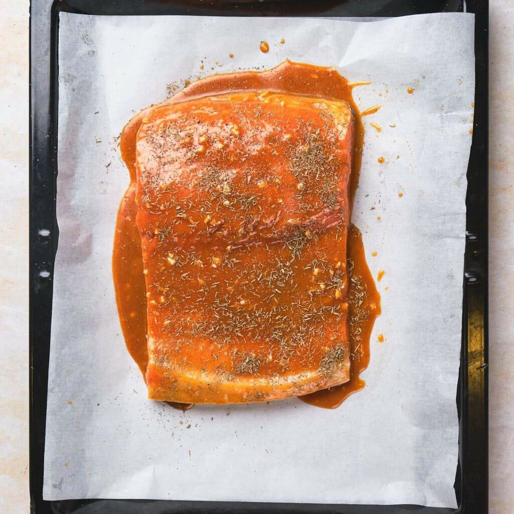 Salmon fillet coated in a red sauce and sprinkled with herbs on a baking sheet lined with parchment paper.