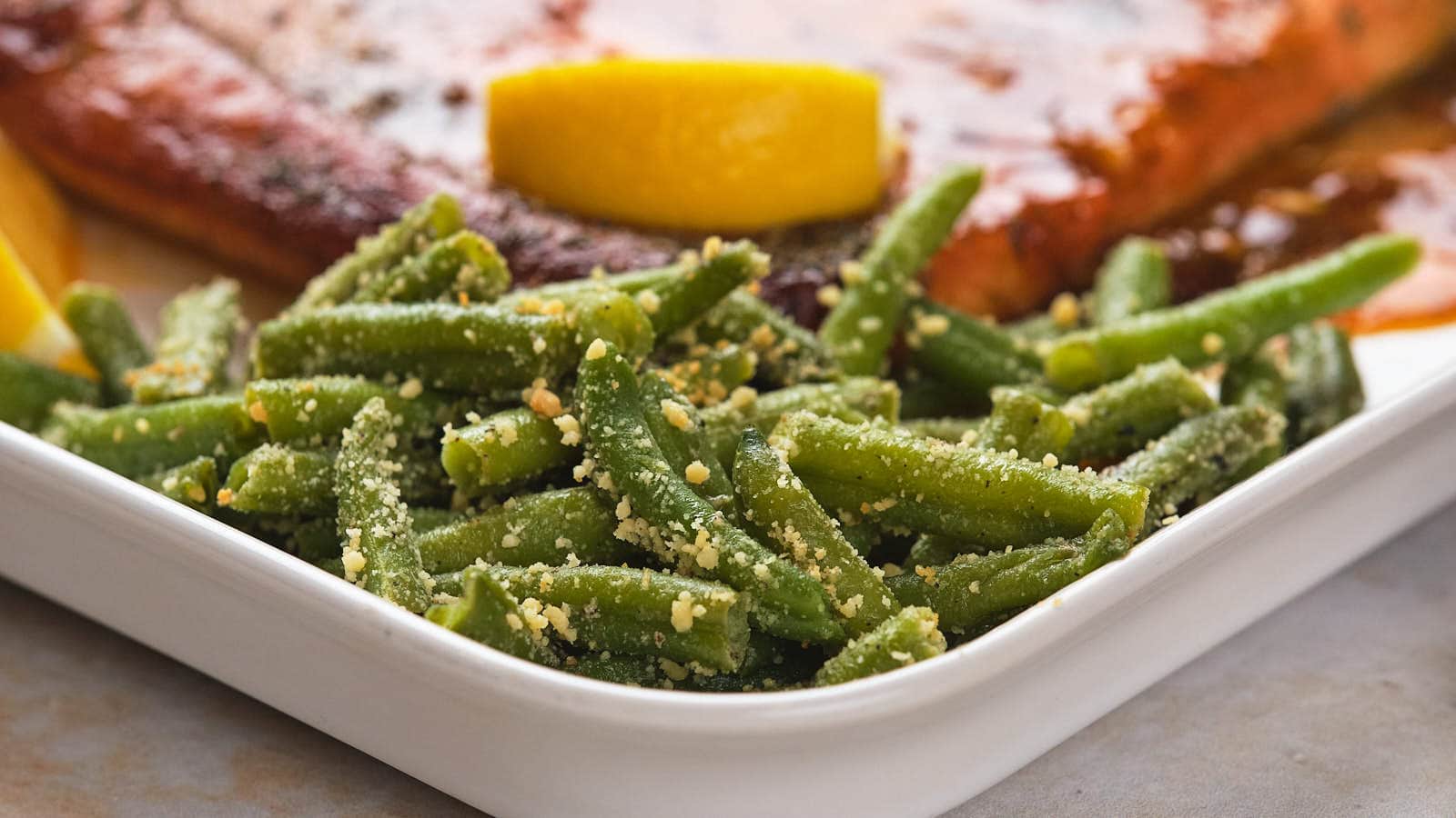 A plate with green beans sprinkled with seasoning and grated cheese, alongside a lemon wedge and a portion of glazed salmon.