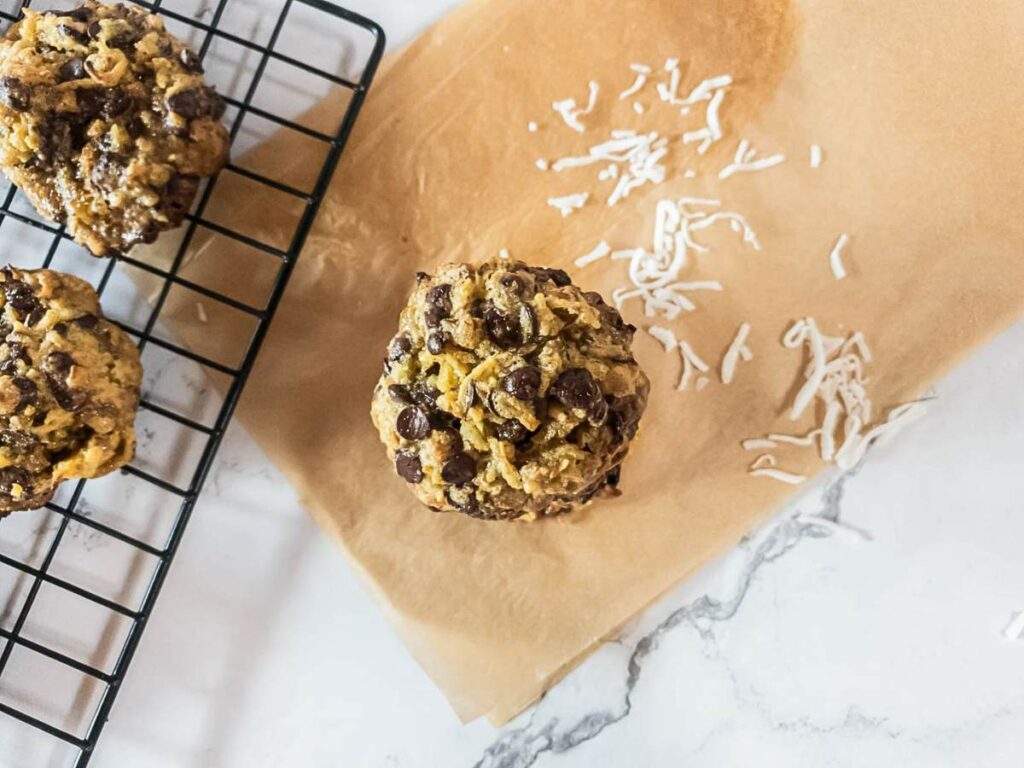 Keto magic cookies on parchment paper, next to a cooling rack, with shredded coconut scattered on the surface.