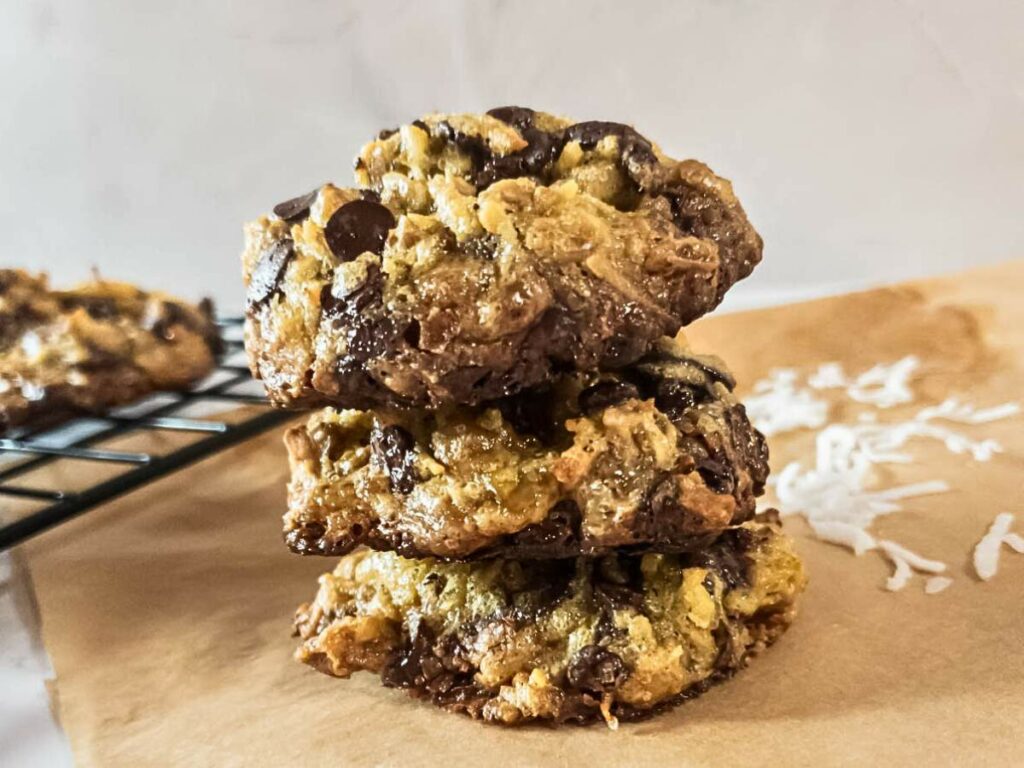 Three keto magic cookies stacked on a piece of parchment paper, with a cooling rack and scattered coconut flakes in the background.
