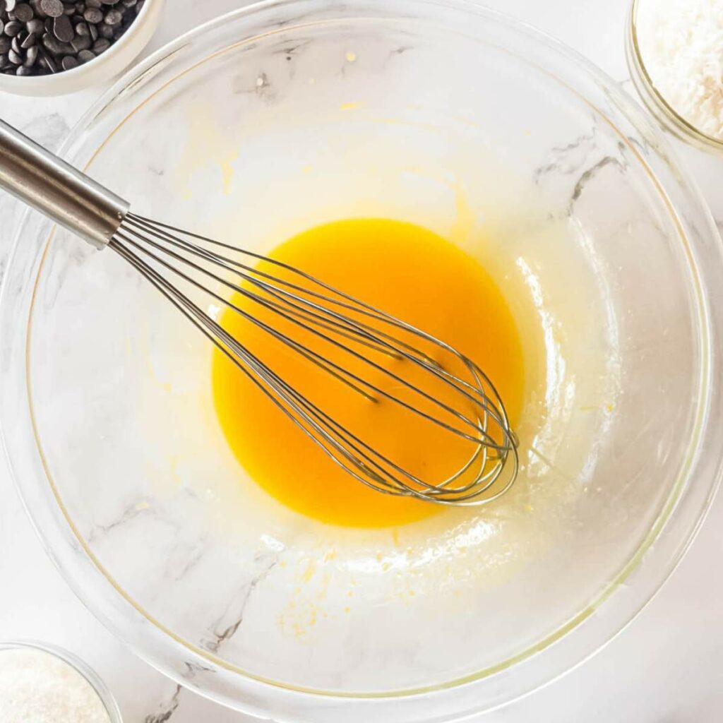 A whisk in a glass bowl with yellow liquid mixture. Nearby are bowls of chocolate chips, flour, and sugar.