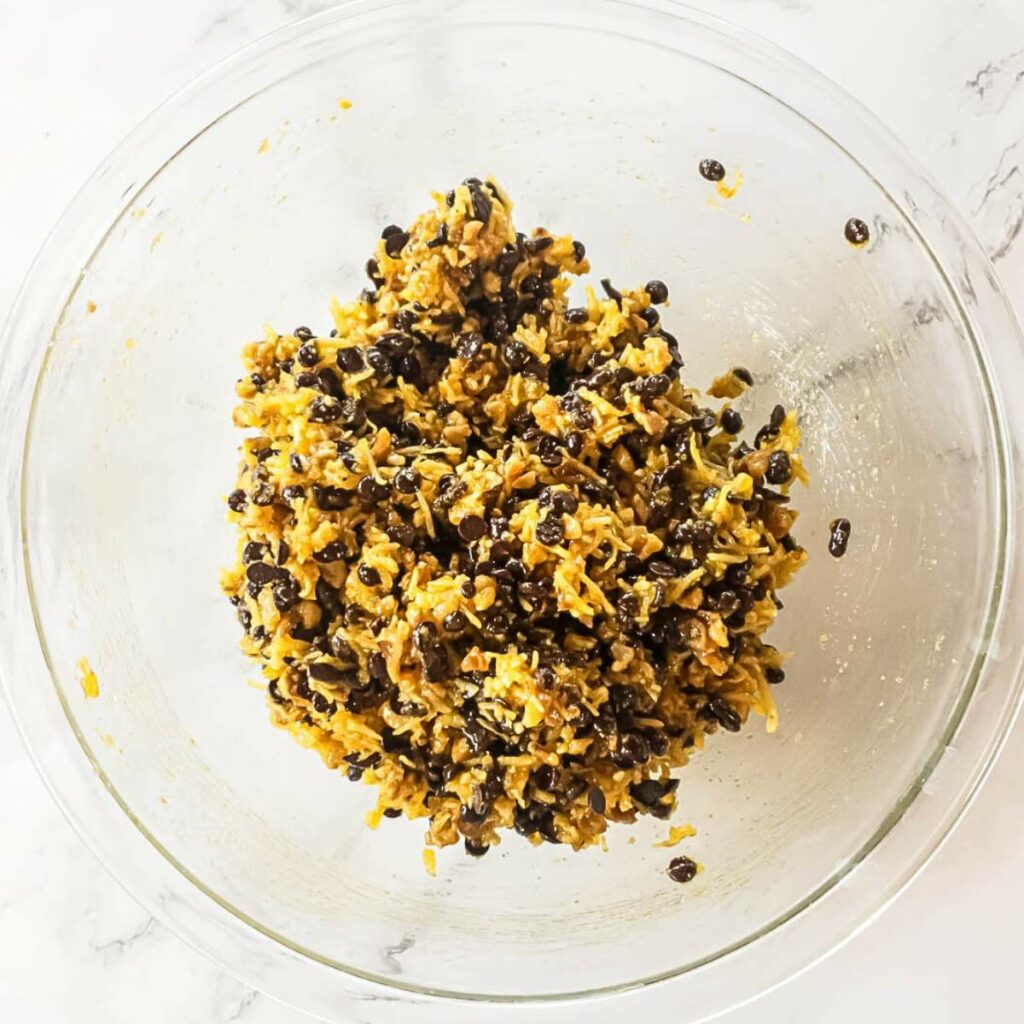 A glass bowl containing a mixture of shredded carrots, chocolate chips, and oats sits on a marbled surface.