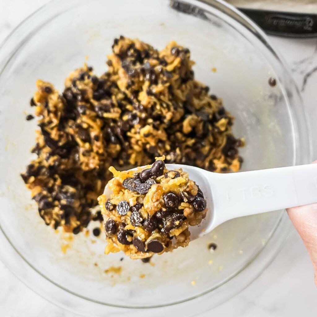Spoon holding a mixture with chocolate chips over a glass bowl filled with the same mixture.