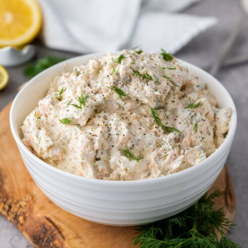 A white bowl filled with salmon dip with cream cheese garnished with dill on a wooden board.