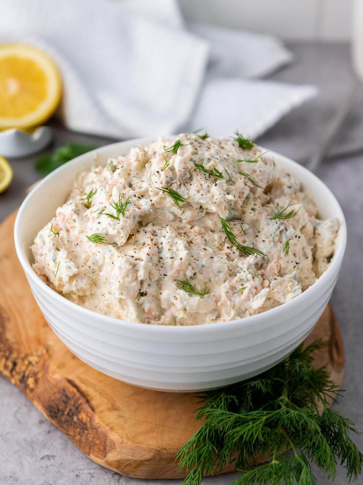 A white bowl filled with salmon dip with cream cheese garnished with dill on a wooden board.