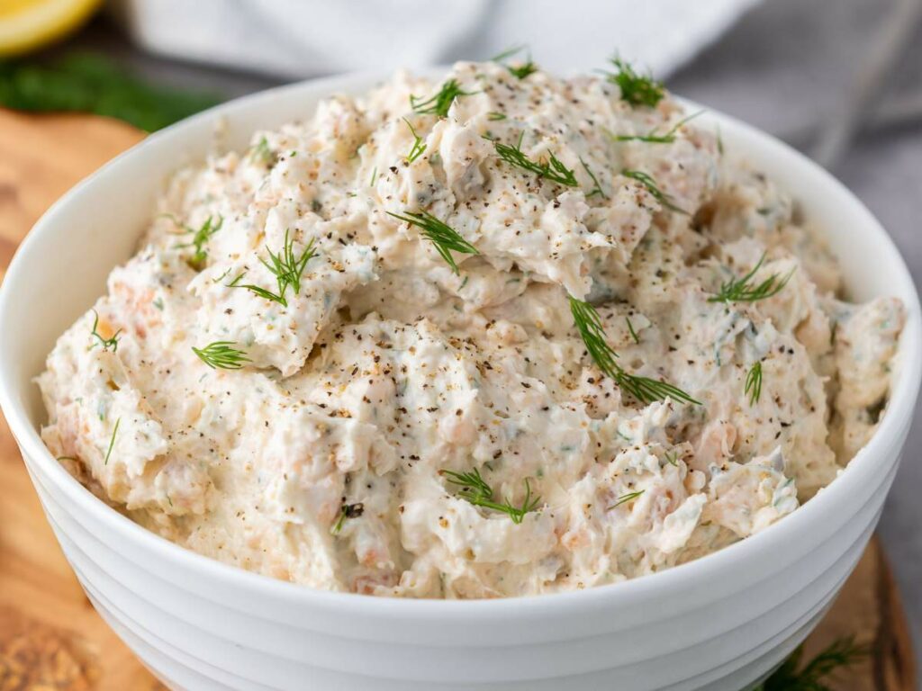 A bowl of salmon dip with cream cheese topped with black pepper and fresh dill, placed on a wooden surface.