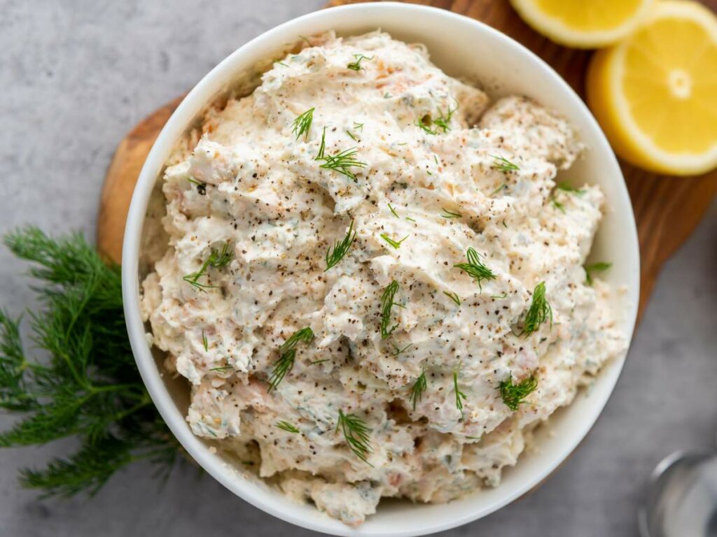 Bowl of salmon dip with cream cheese garnished with dill and pepper, placed on a wooden board.