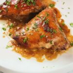 Two pieces of glazed oven-baked chicken garnished with chopped herbs sit on a plate with apricot sauce. Text reads "Delicious Oven-Baked Apricot Chicken.