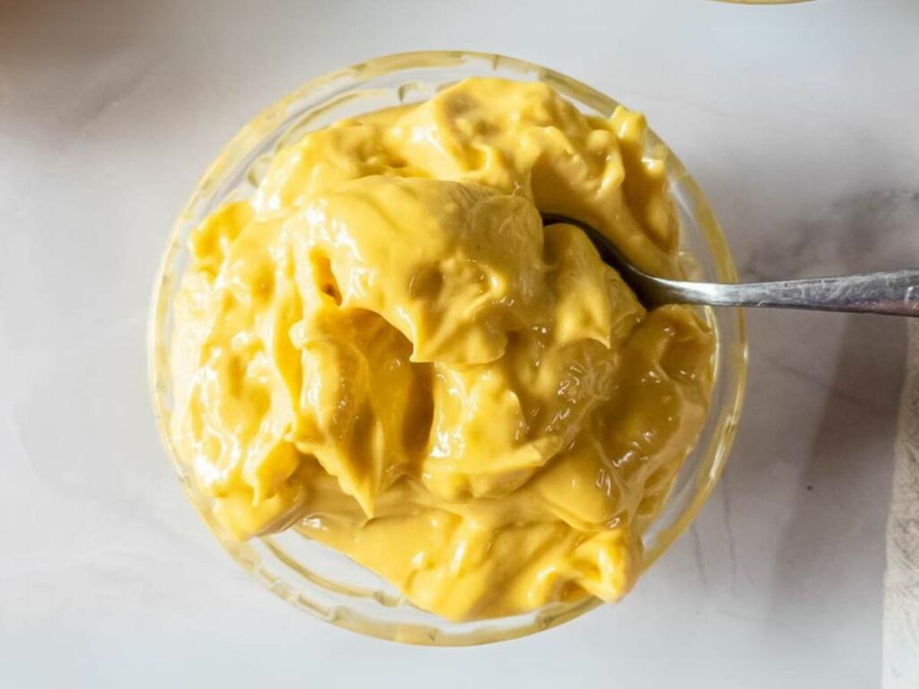A glass bowl filled with thick, yellow mayonnaise with a spoon resting in it, viewed from above.