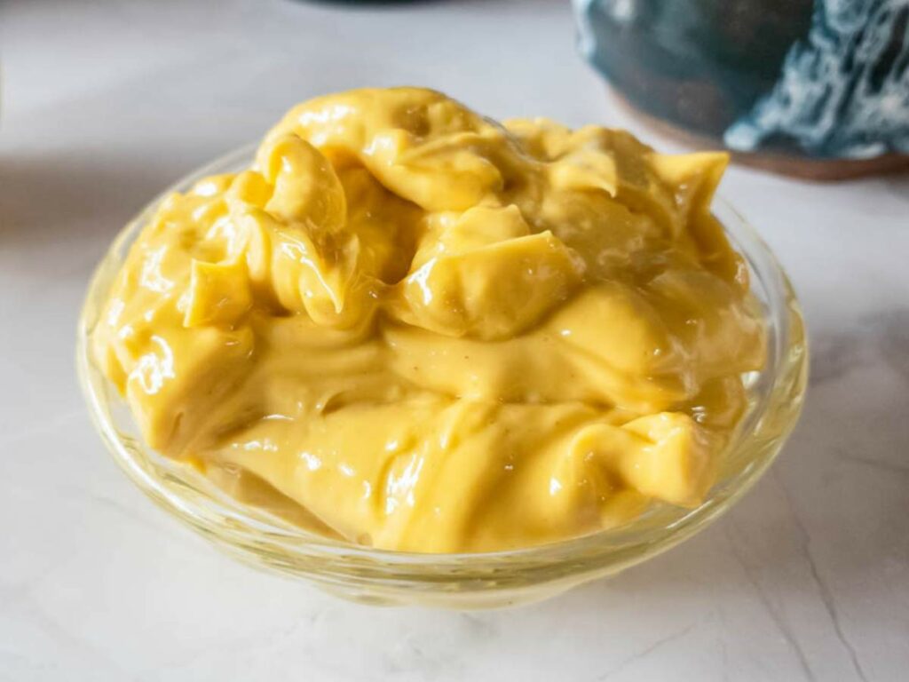 A small clear glass bowl filled with thick, yellow mayonnaise sits on a white surface.